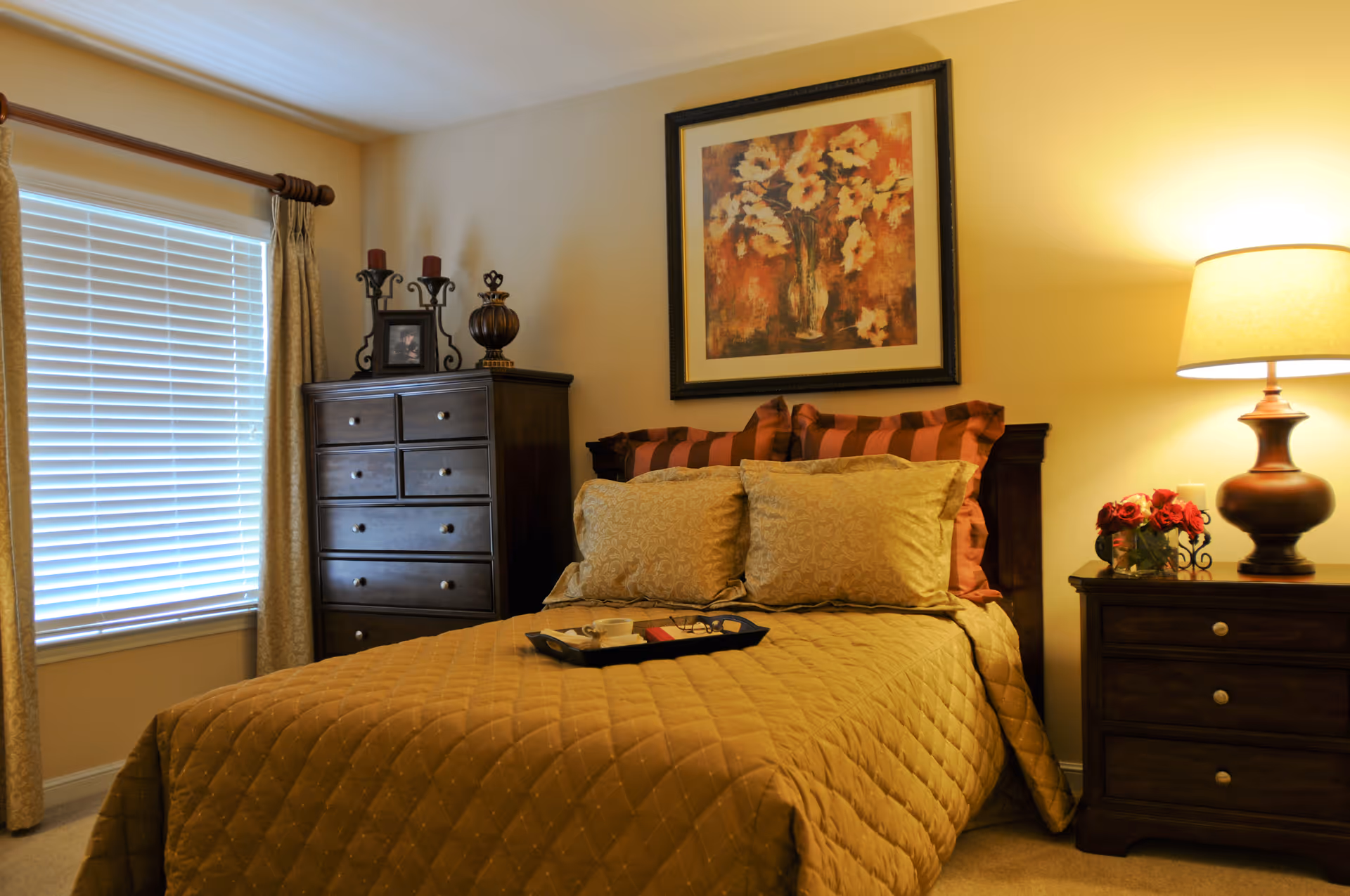 A cozy bedroom with a neatly made bed featuring a golden quilt and multiple pillows. There is a dark wooden dresser with decorative items and a framed picture on the wall above the bed. A bedside table with a lamp and a small flower arrangement is on the right side. A window with closed blinds and curtains is on the left side of the room.