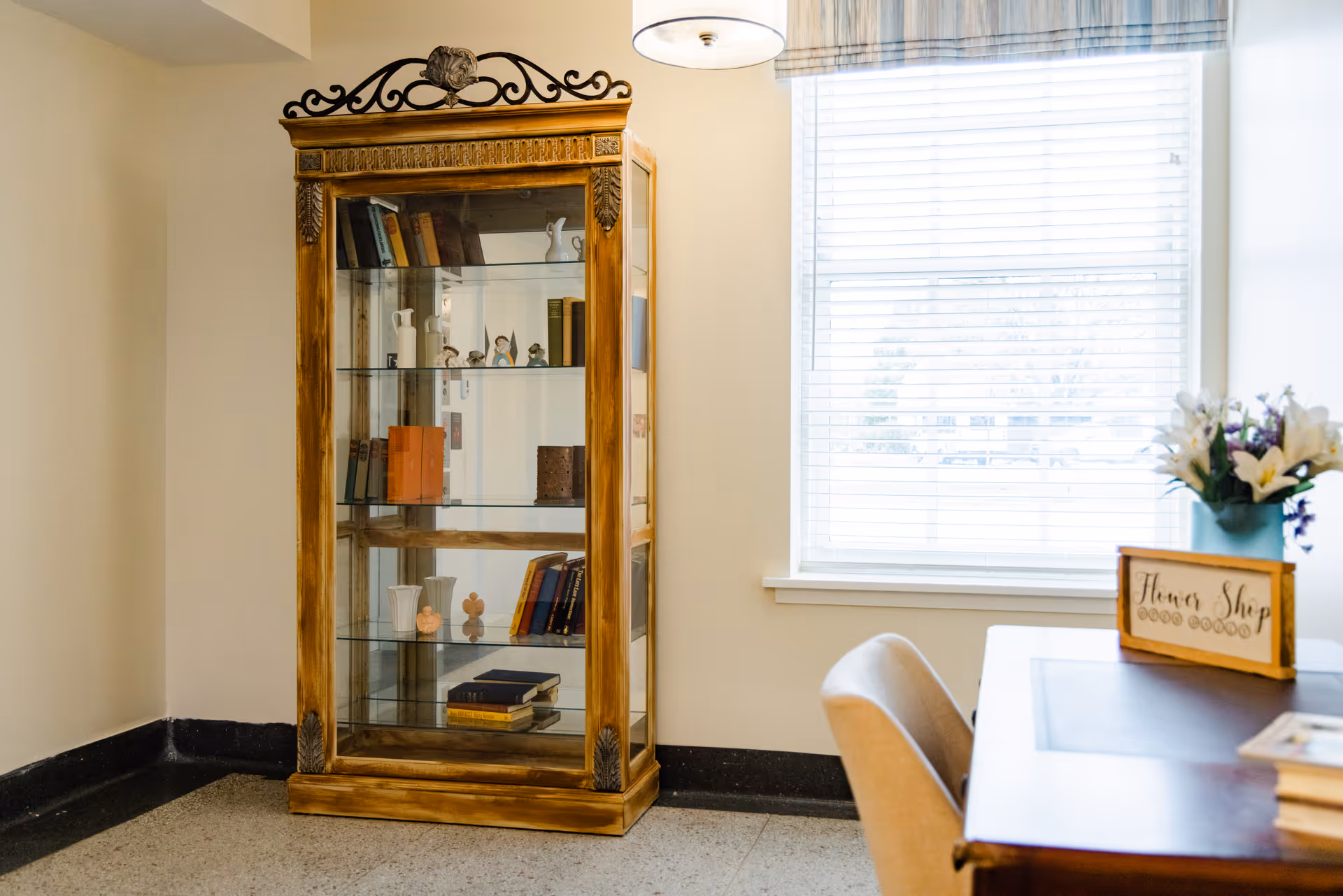 A cozy interior corner featuring a wooden display cabinet with glass shelves holding books and decorative items, next to a window with blinds. A desk with a chair is partially visible, adorned with a flower arrangement and a small sign that reads 'Flower Shop'.