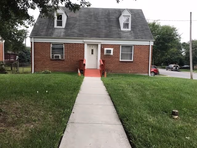 Front view of a small brick house with a gray shingled roof and two dormer windows. A concrete walkway leads to a white front door with red steps and railings. The house has two windows on the front, one with an air conditioning unit. There is green grass on both sides of the walkway and trees in the background.