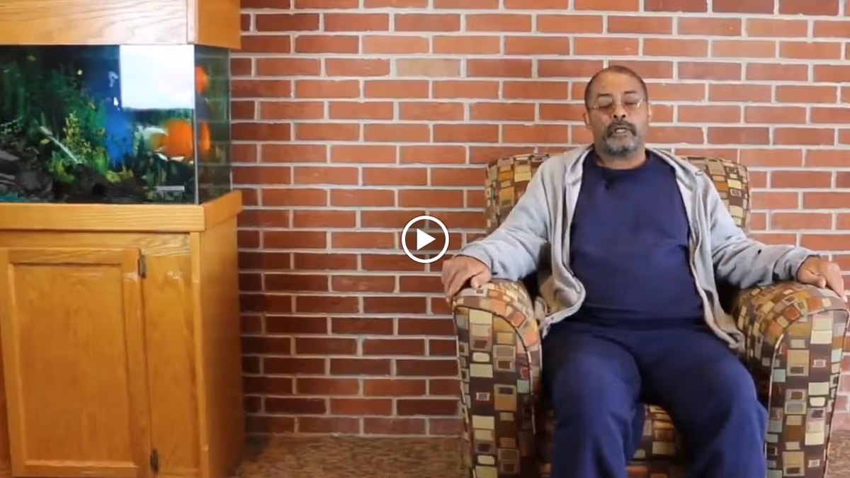 An elderly man wearing glasses, a navy blue shirt, and a light gray jacket sits in a patterned armchair against a red brick wall. To his left, there is a wooden cabinet with a large fish tank on top containing colorful fish and aquatic plants.