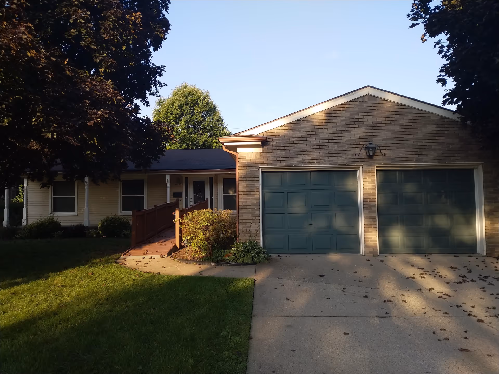 Exterior view of a single-story residential building with a two-car garage featuring green doors. There is a concrete driveway and a wooden ramp leading to the front door. The building is surrounded by green grass and trees with some shadows cast on the driveway and garage doors.