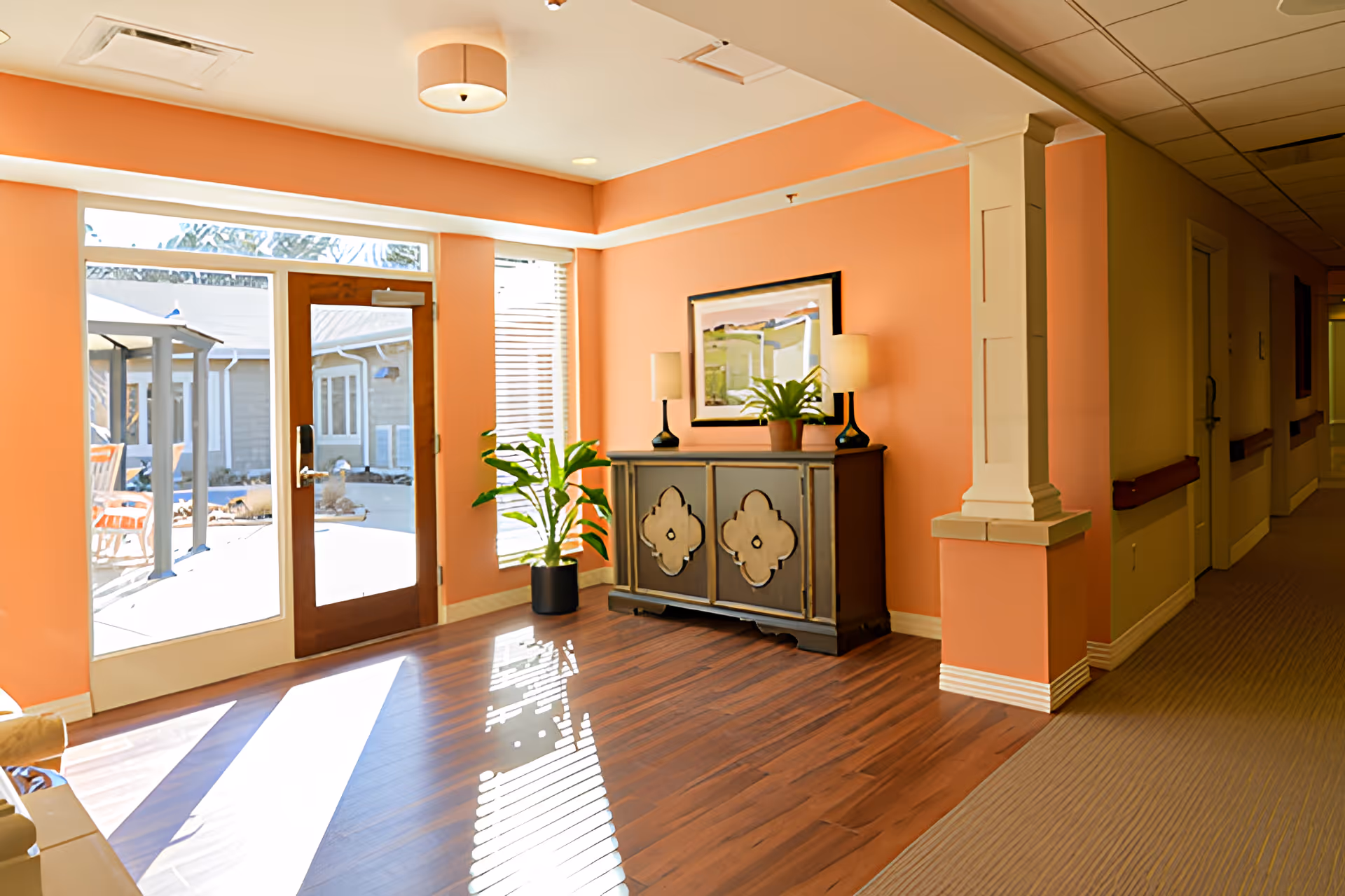 Sunlit lobby area with glass doors to an outdoor courtyard, a decorative cabinet, potted plant, and coral-colored walls.