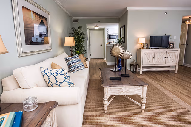 A cozy living room with a beige sofa adorned with patterned pillows, a wooden coffee table with decorative items, a side table with a lamp, a framed picture on the wall, a TV on a wooden cabinet, and a view into a small kitchen area in the background.