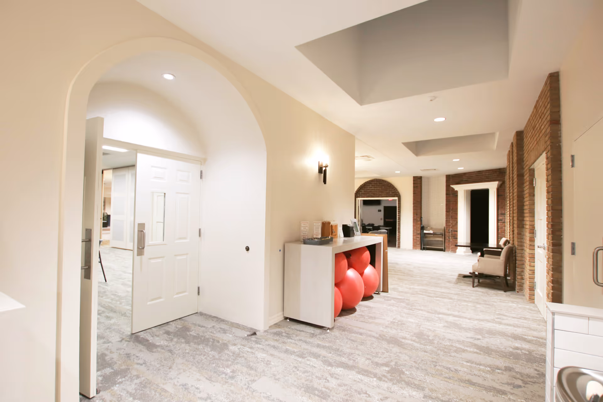 A bright hallway in a senior living facility with light-colored walls and carpeted floor. The hallway features an arched doorway with a white door partially open, a small table holding red exercise balls underneath, and seating areas with chairs along the brick wall. The ceiling has recessed lighting and decorative architectural details.