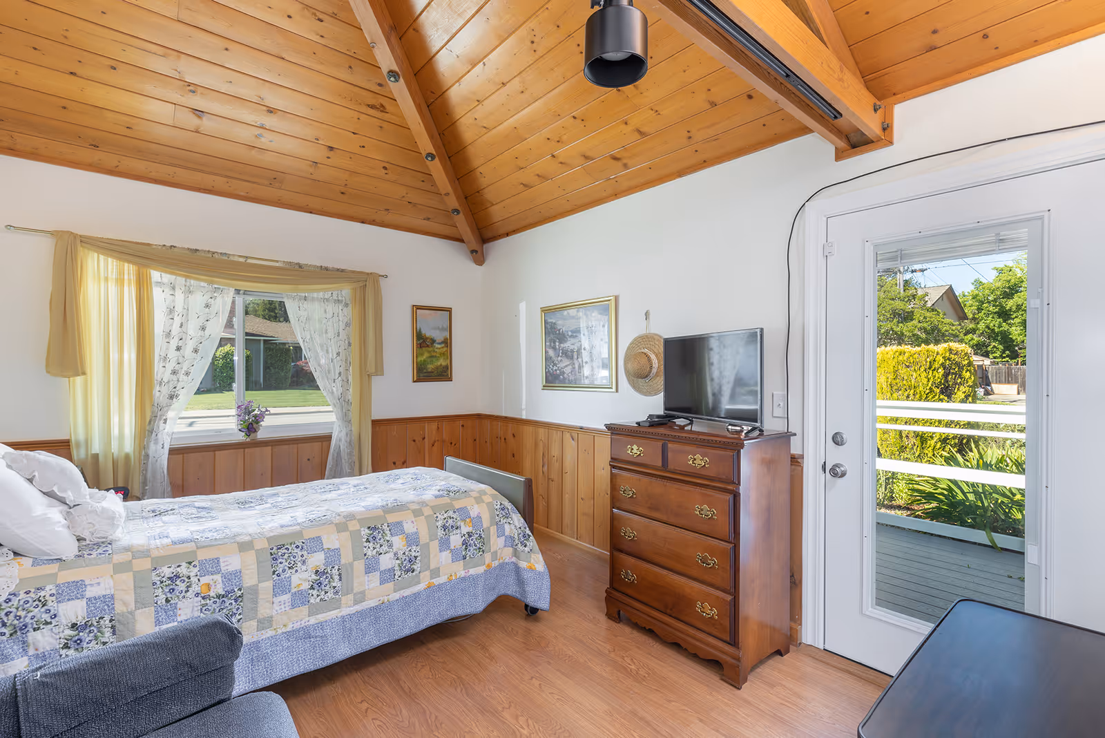 A cozy bedroom with a wooden vaulted ceiling and wood paneling on the lower half of the walls. The room features a bed with a patchwork quilt, a window with sheer and yellow curtains, a wooden dresser with a flat-screen TV on top, framed artwork on the walls, and a glass door leading outside to a porch with greenery visible.
