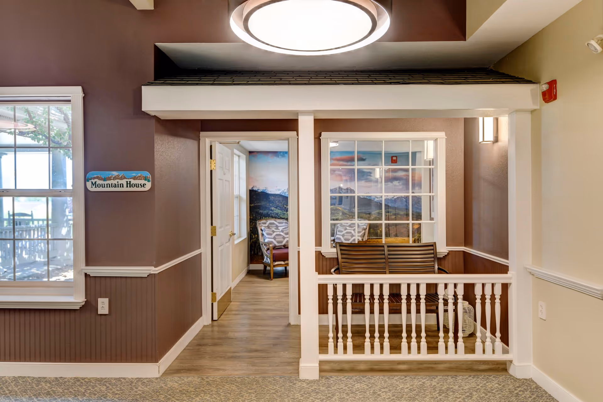 Interior view of a senior living facility hallway with a small porch-like seating area featuring a wooden bench and two cushioned chairs. The walls are painted brown and beige, with a sign that reads 'Mountain House'. There is a large round ceiling light above and a window showing an outdoor area with trees.