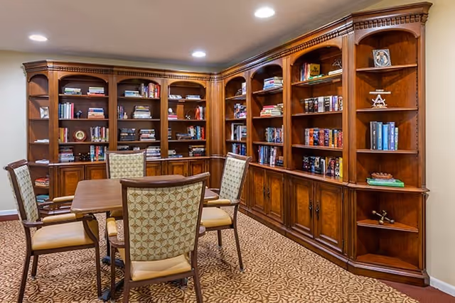 A cozy library room with wooden bookshelves filled with books and decorative items. There is a wooden table in the center surrounded by four upholstered chairs with patterned fabric. The room has recessed ceiling lights and a patterned carpet.