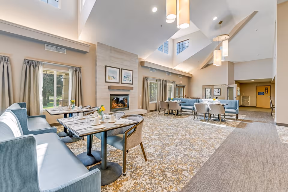 A bright and spacious dining area in an assisted living facility with large windows, high ceilings, and modern pendant lights. The room features several dining tables set with white dishes and silverware, blue cushioned bench seating, and beige chairs. A fireplace with two framed pictures above it is centered on one wall, and the carpet has a patterned design.