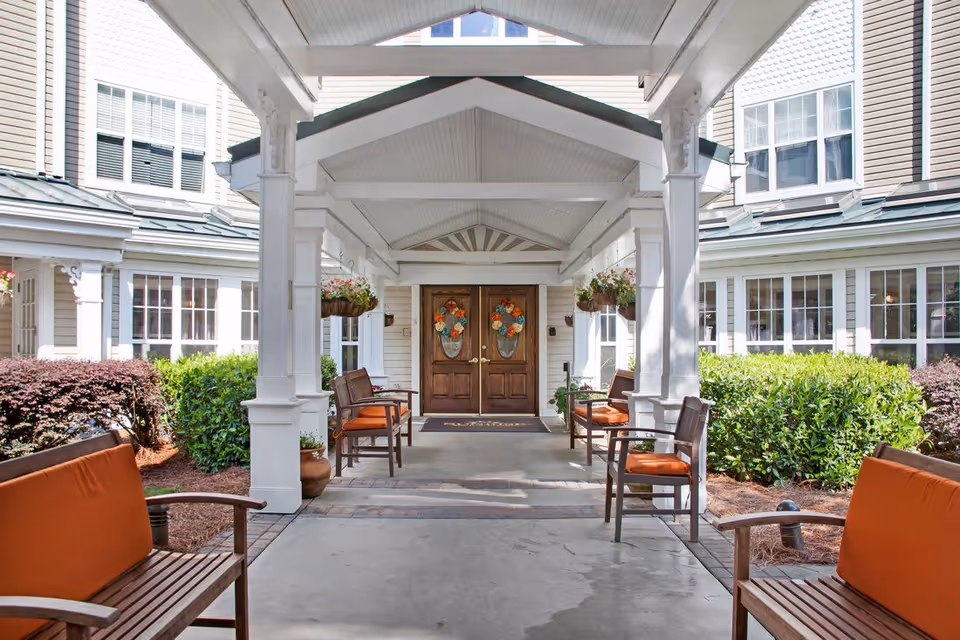 Covered front entrance with double wooden doors decorated with wreaths, benches with orange cushions, hanging planters, and surrounding shrubs.