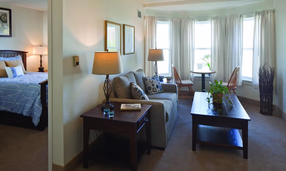 A bright living room with a sofa, coffee table, side table lamps and a small dining table by bay windows, with a bedroom visible through an open doorway.