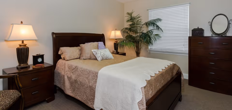 A neatly made bed with a beige patterned bedspread and white throw blanket in a bedroom. The room features two bedside tables with lamps, a wooden dresser with a round mirror, a potted plant near a window with closed blinds, and a chair with a leopard print cushion.