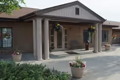 Senior living facility entrance with brick facade, columns, and potted plants along walkway.