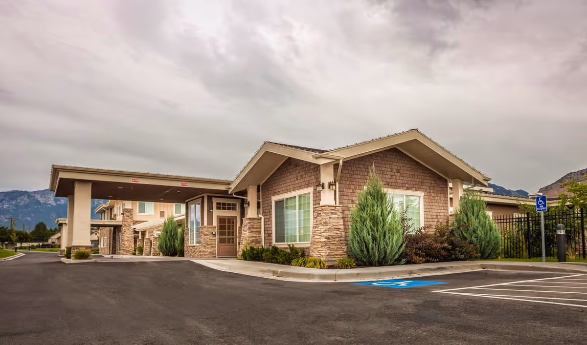 Exterior view of Ashford Assisted Living & Memory Care facility showing a single-story building with stone and wood siding, large windows, and a covered entrance. There are landscaped bushes and trees around the building, a parking lot with handicap parking spaces, and mountains visible in the background under a cloudy sky.