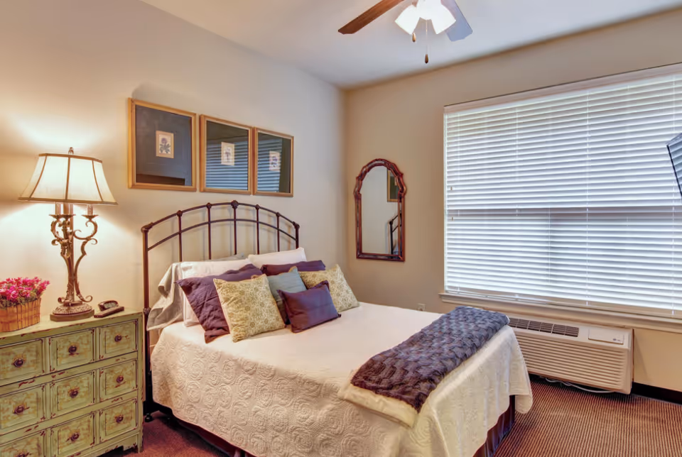 A cozy bedroom with a bed featuring multiple pillows and a white quilted bedspread. To the left of the bed is a green distressed nightstand with a decorative lamp and a basket of flowers. Above the bed are three framed pictures, and a mirror hangs on the adjacent wall. A large window with closed blinds is on the right side, and a ceiling fan with a light is visible on the ceiling.