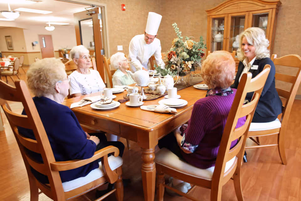 A group of elderly women seated around a wooden dining table set with plates, cups, and napkins. A chef in a white uniform and hat is serving food, while a woman in a black blazer smiles and interacts with the group. The room has wooden flooring, a china cabinet with dishes, and a floral centerpiece on the table.