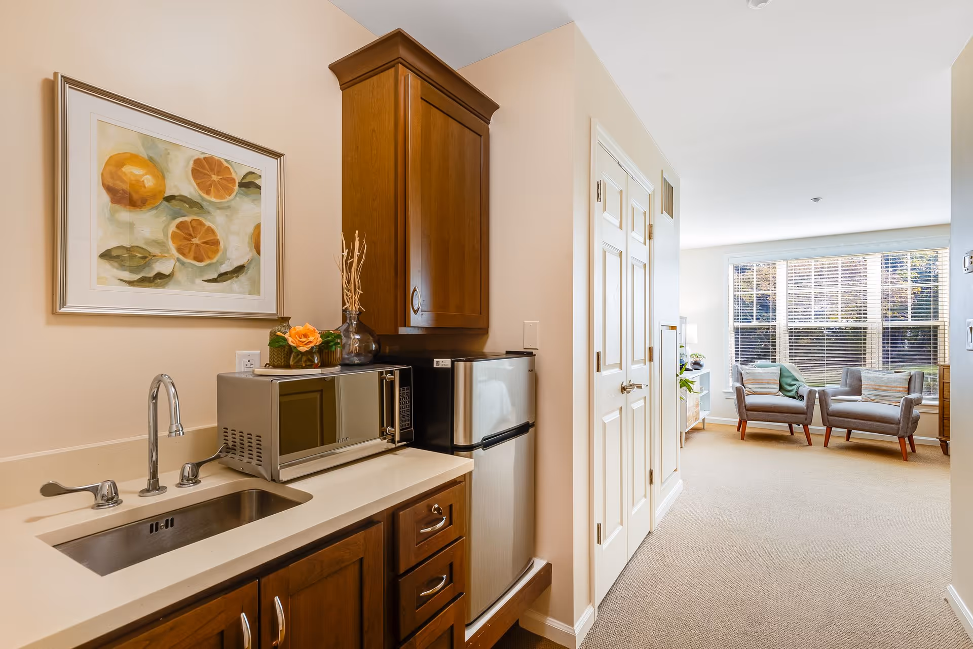 A small kitchenette area with a stainless steel sink, microwave, and mini refrigerator. Above the microwave is a wooden cabinet, and a framed painting of oranges hangs on the wall. In the background, there is a bright living room with two armchairs near a large window with blinds.