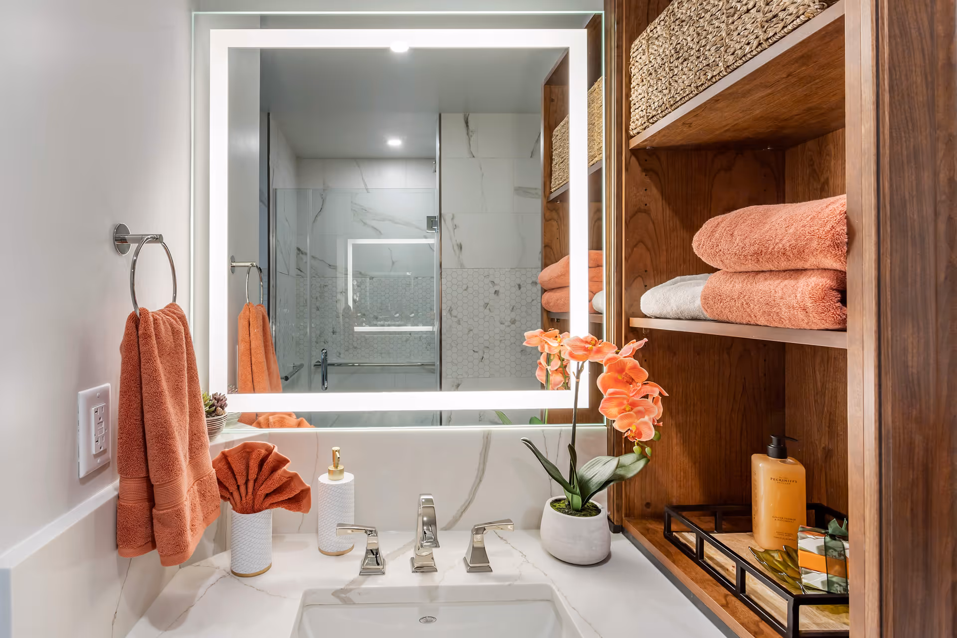 A modern bathroom vanity with a white marble countertop, a rectangular sink, and a large illuminated mirror. On the countertop are a soap dispenser, a folded orange towel in a holder, and a potted orange orchid. To the right, wooden shelves hold folded orange and white towels, woven baskets, and a tray with a lotion bottle and soap. A glass shower with marble and hexagonal tile walls is visible in the mirror's reflection.