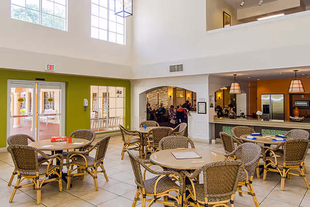 Bright communal dining area with round tables and wicker chairs, a serving counter and high windows.