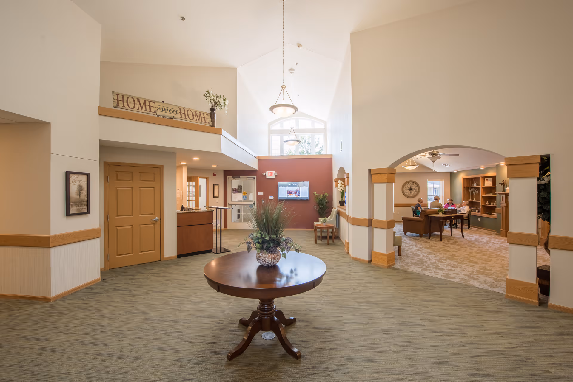 A spacious and well-lit common area in a senior living facility with a round wooden table and a decorative plant in the center. The room features high ceilings, beige walls with wooden trim, and a sign that reads 'HOME sweet HOME' above a doorway. To the right, there is an arched opening leading to a sitting area where several elderly people are seated around a table. The area has carpeted floors and warm lighting.