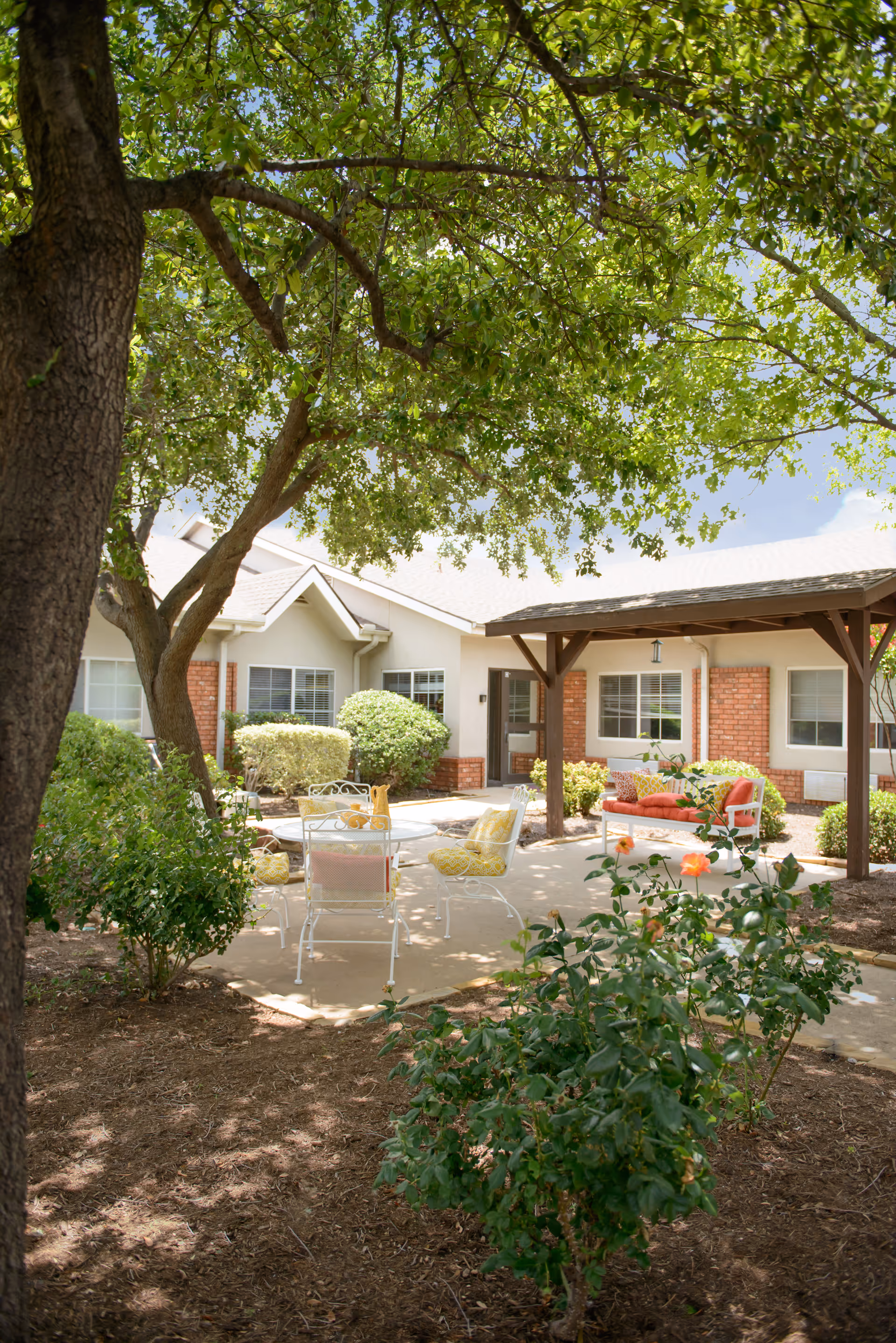Outdoor patio area at a senior living facility with a large tree providing shade, white metal chairs and a table with yellow cushions, a red cushioned bench under a wooden pergola, surrounded by bushes and flowers in a garden setting.