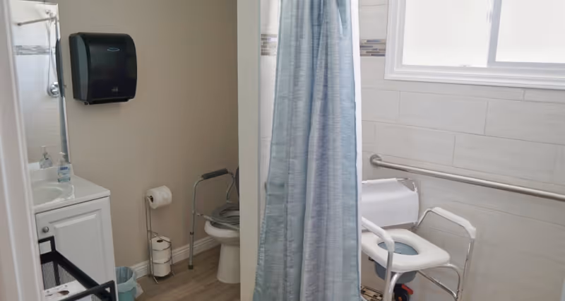 A bathroom with a toilet equipped with safety rails and a shower chair behind a blue shower curtain. There is a sink with a soap dispenser, a paper towel dispenser mounted on the wall, and a toilet paper holder with extra rolls. The walls are tiled and there is a window letting in natural light.
