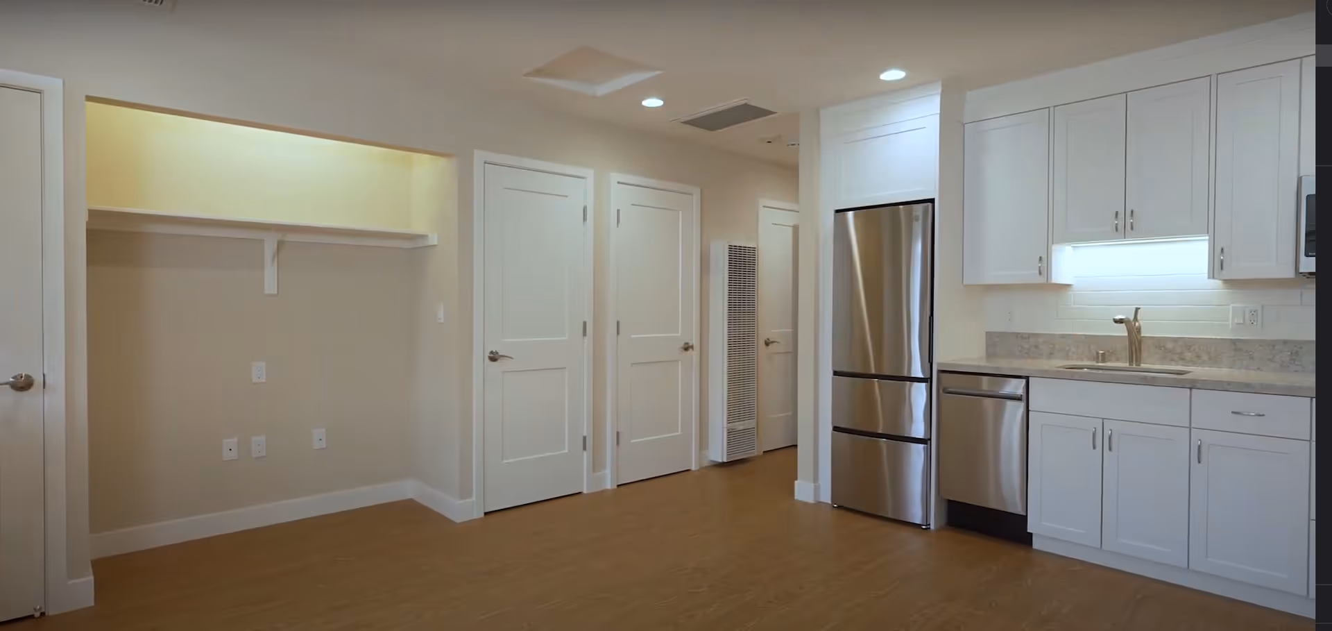 Interior view of a modern kitchen area with white cabinets, a stainless steel refrigerator, dishwasher, and a sink with a faucet. There are three closed white doors and a small alcove with a shelf and electrical outlets on the left side. The floor is wood, and the walls are painted beige.