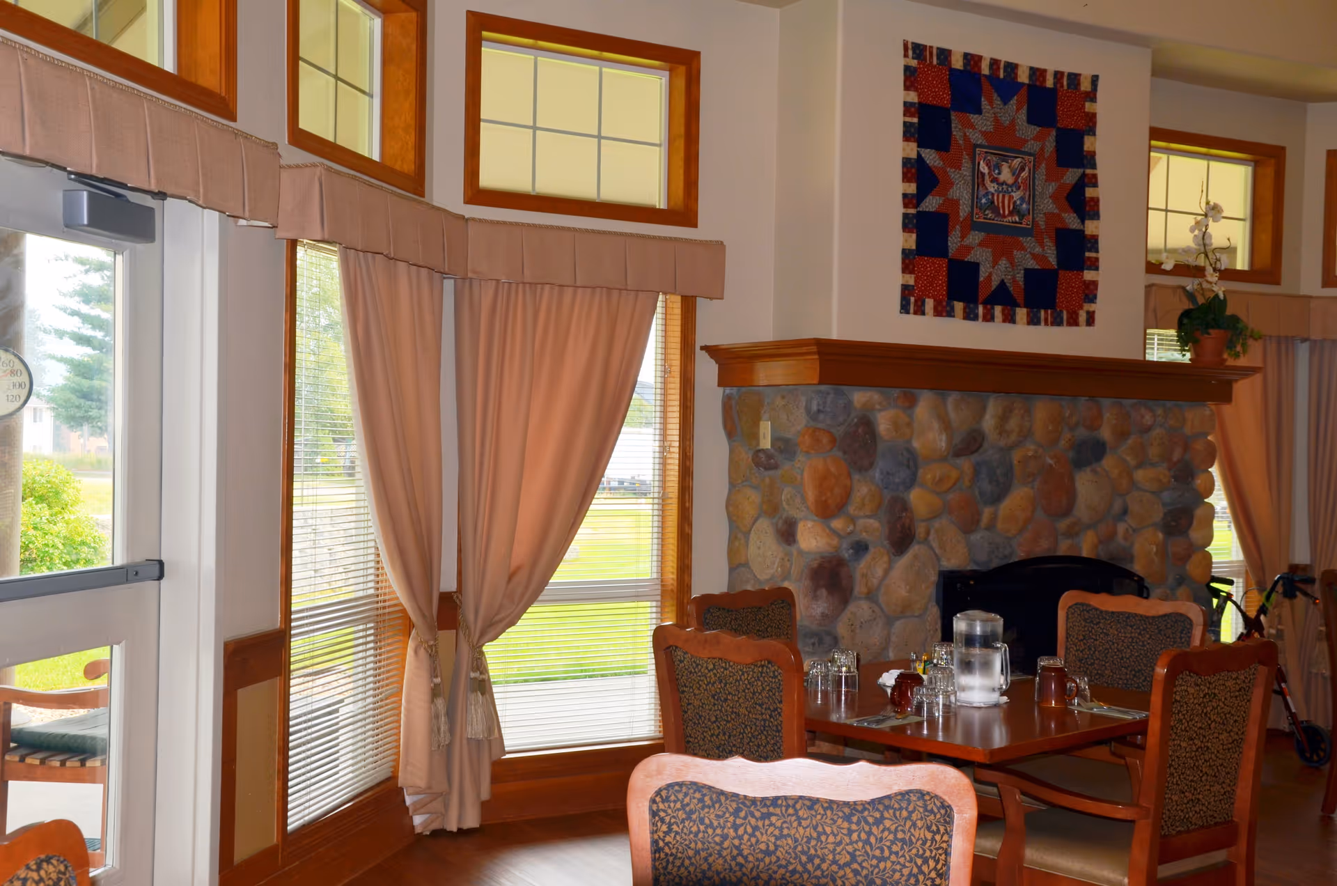 A cozy interior dining area with a wooden table set for four, featuring a water pitcher and glasses. The room has large windows with beige curtains and a stone fireplace with a wooden mantle. Above the fireplace hangs a colorful quilt with a patriotic design. The space is warmly lit with natural light coming through the windows.