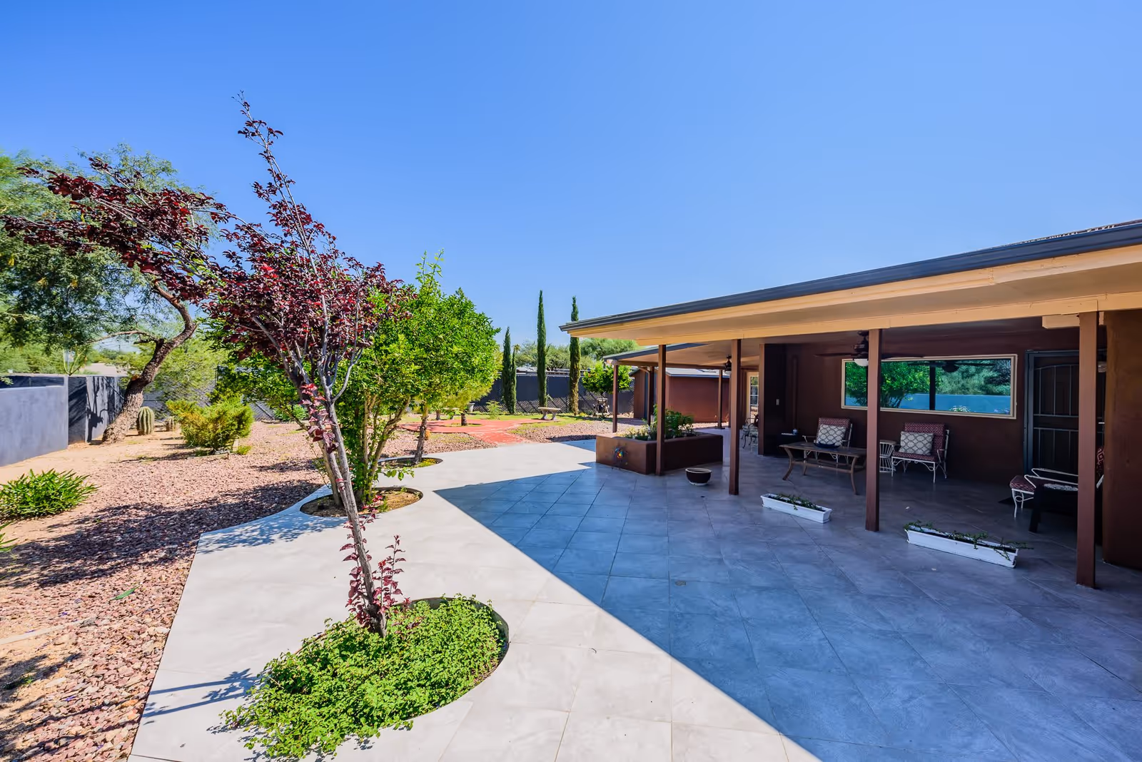 Outdoor patio area at Oak Haven Assisted Living at Casas Adobes with tiled flooring, seating arrangements under a covered porch, and landscaped garden with trees and shrubs under a clear blue sky.