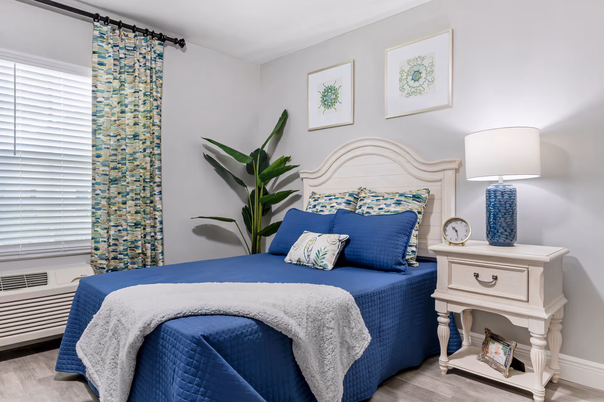 A bedroom with a bed covered in a blue quilt and decorative pillows, a white wooden headboard, a white nightstand with a blue lamp and a clock, two framed botanical prints on the wall, a large green plant in the corner, and a window with patterned curtains and blinds.