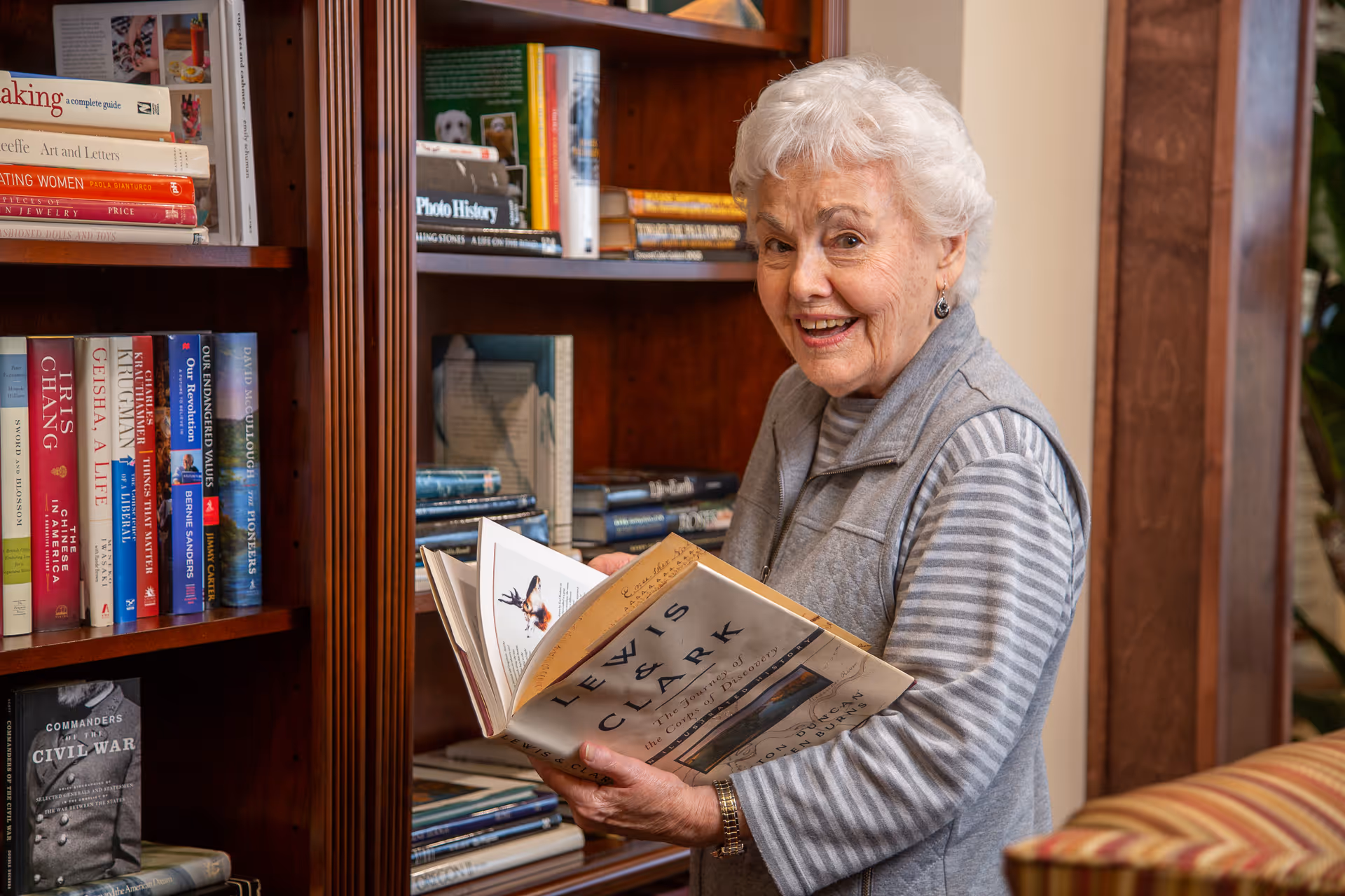 An elderly woman with white hair wearing a gray striped shirt and gray vest is standing in front of a wooden bookshelf filled with books. She is holding an open book titled 'Lewis & Clark' and smiling at the camera. The setting appears to be a cozy indoor area with warm lighting.