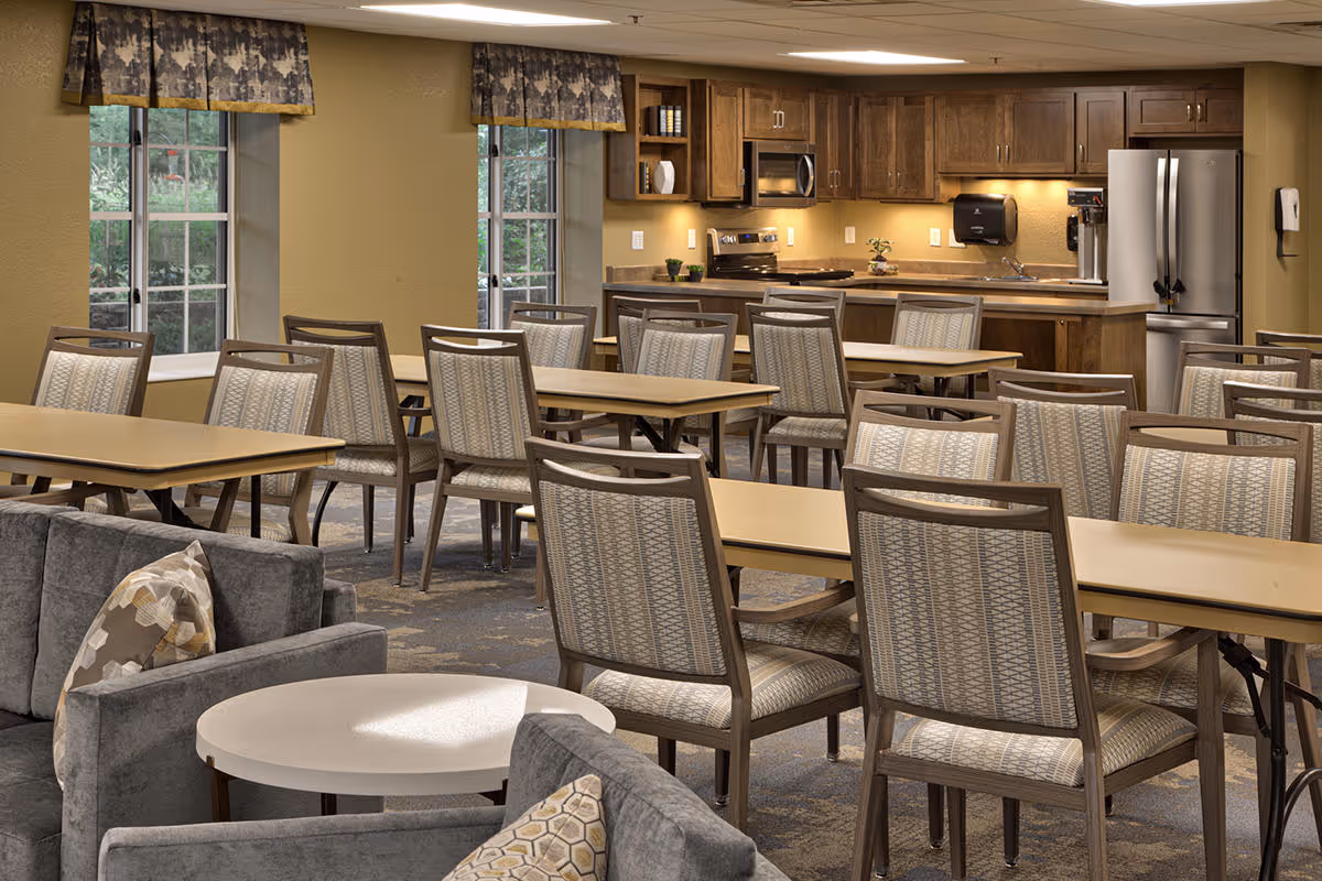A communal dining area with multiple tables and chairs arranged in rows. In the background, there is a kitchen area with wooden cabinets, a stainless steel refrigerator, microwave, stove, and a sink. The room has two windows with patterned valances and a carpeted floor. In the foreground, there are gray upholstered sofas with patterned pillows and a round white table.