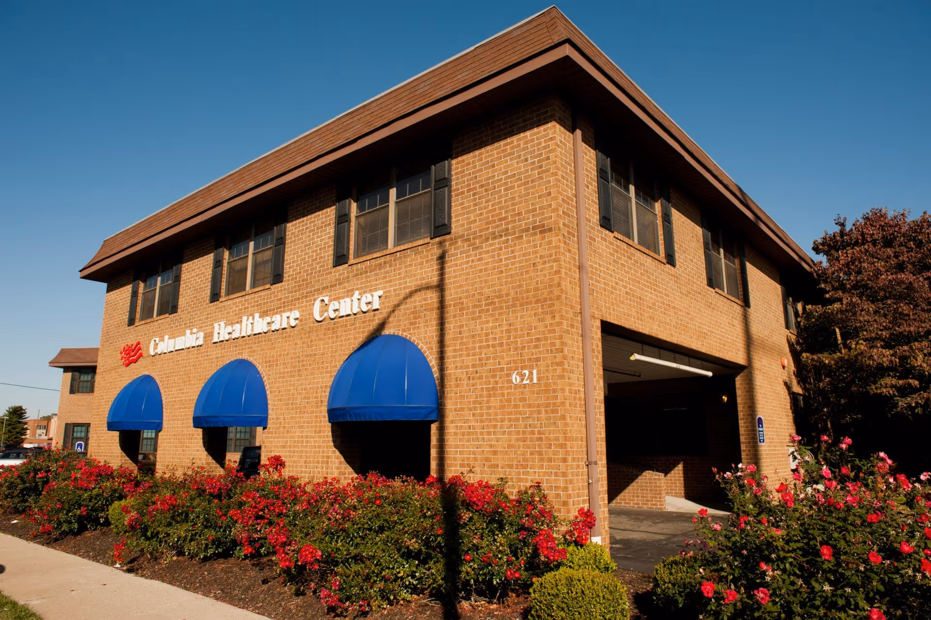Exterior view of Columbia Healthcare Center, a two-story brick building with blue awnings over the windows and a garden with red flowers in front. The building number 621 is visible on the corner.