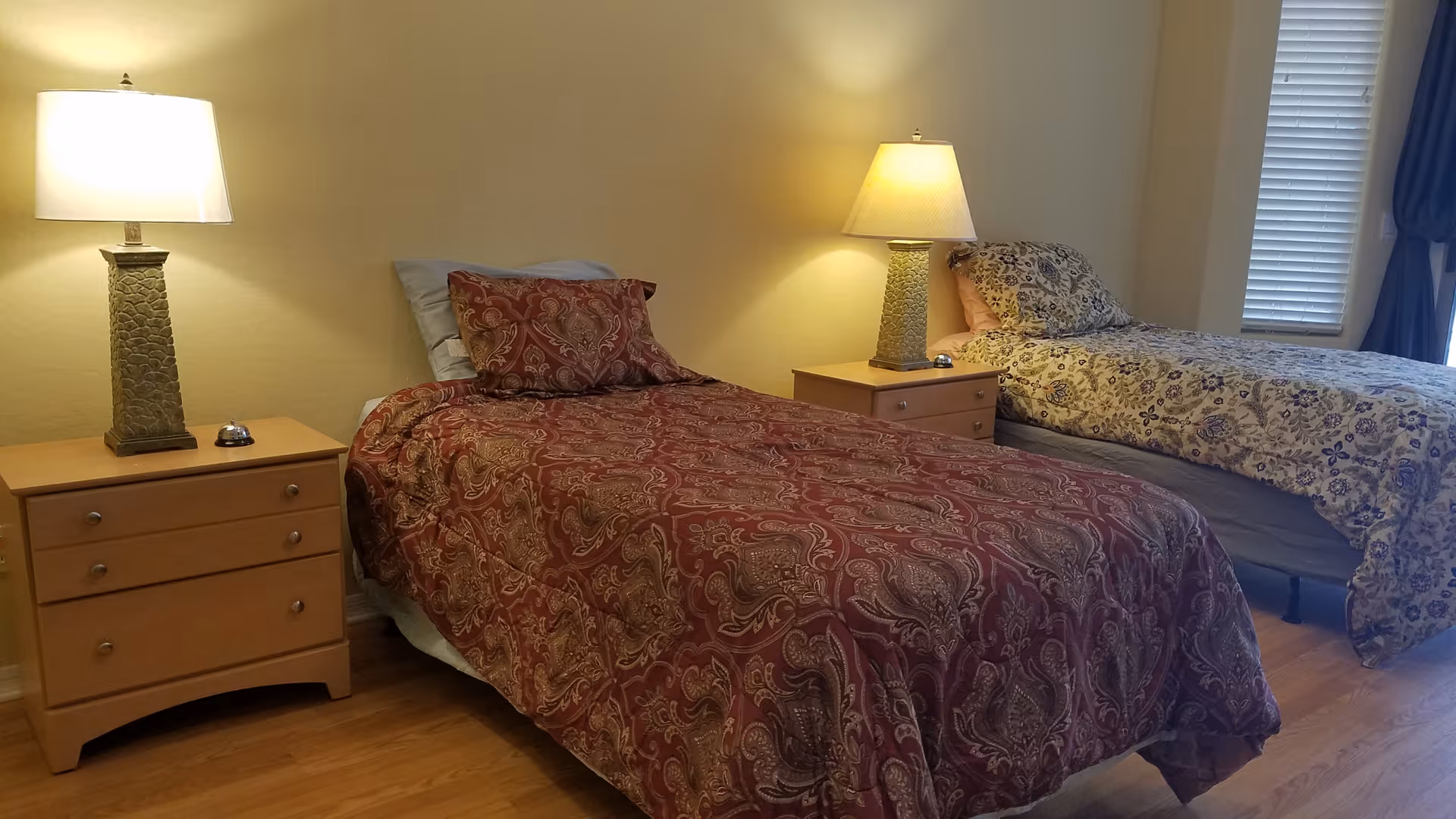 A bedroom with two single beds, each with patterned bedspreads and pillows. Between the beds are two wooden nightstands, each with a lamp and a small bell. The room has wooden flooring and a window with blinds and curtains on the right side.