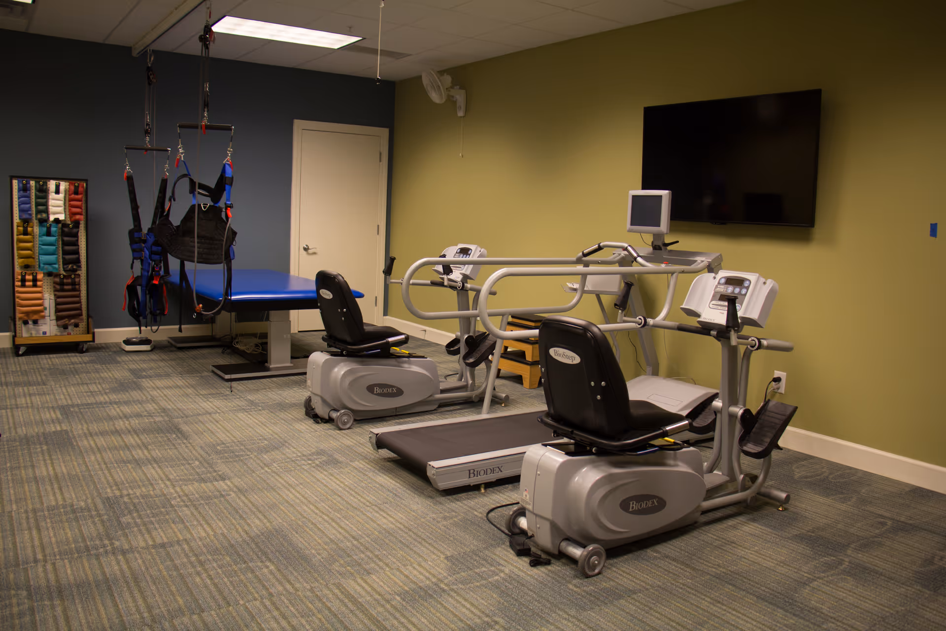 Therapy/exercise room with recumbent bikes, a treadmill, a therapy table with suspension harnesses, and a wall-mounted TV.