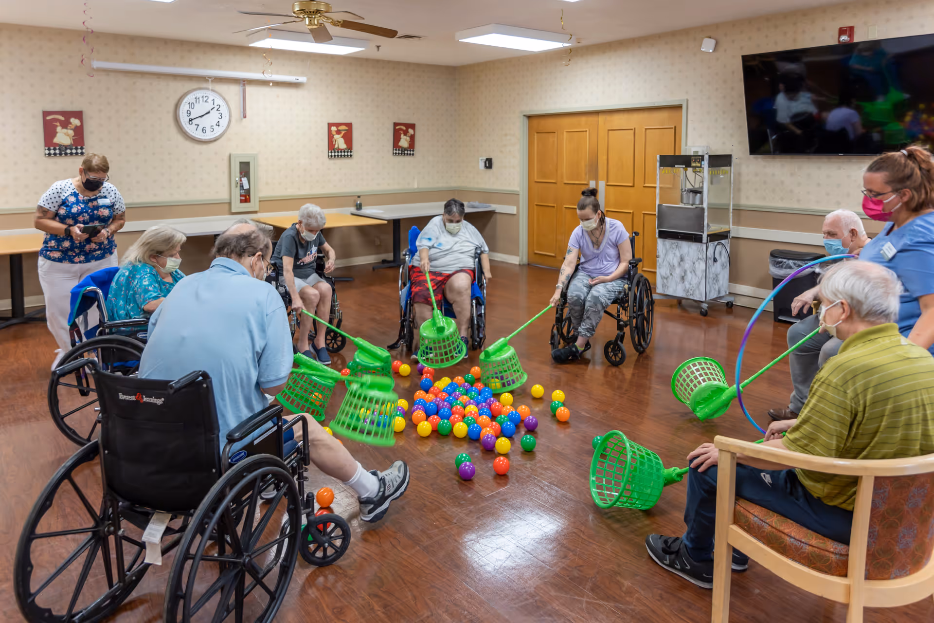 A group of elderly individuals, some in wheelchairs, participating in a seated activity involving green baskets and colorful plastic balls in a spacious room. Two caregivers wearing masks are assisting them. The room has wooden floors, beige wallpaper, a wall clock, a ceiling fan, and a large TV mounted on the wall.