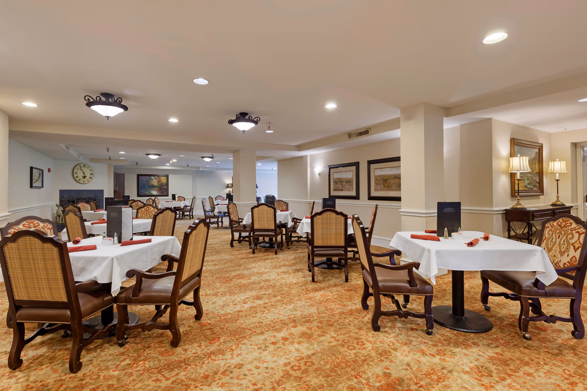 A spacious dining room with multiple tables covered in white tablecloths and set with red napkins. The room features ornate wooden chairs with cushioned seats and backs, warm lighting from ceiling fixtures and lamps, framed artwork on the walls, and a patterned carpet in shades of orange and beige.