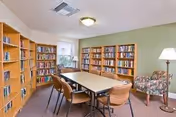 A cozy reading or meeting room with wooden bookshelves filled with books along two walls, a rectangular table surrounded by six chairs in the center, a floral upholstered armchair, a floor lamp, and a window letting in natural light.