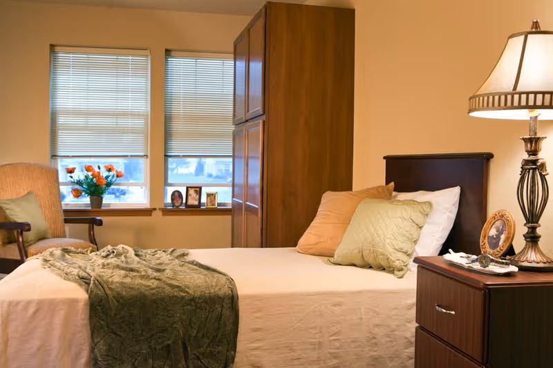 A cozy bedroom with a neatly made bed featuring beige and green pillows and a green throw blanket. There is a wooden nightstand with a decorative lamp and a framed photo. In the background, two windows with blinds partially open show some outdoor scenery. A wooden wardrobe and a cushioned armchair with a green pillow are also visible.