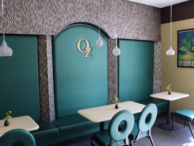 Dining area with green cushioned bench seating along the wall, three small tables with light-colored tops, green chairs with circular backs, small vases with yellow flowers on each table, hanging pendant lights with crystal-like shades, and a decorative wall featuring the letters 'OZ'.