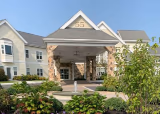 Entrance of a senior living facility named The Knolls, A Bethel Community, featuring a covered driveway with stone pillars and the number 55 displayed at the top. The building is light-colored with multiple windows and surrounded by landscaped greenery and plants under a clear blue sky.