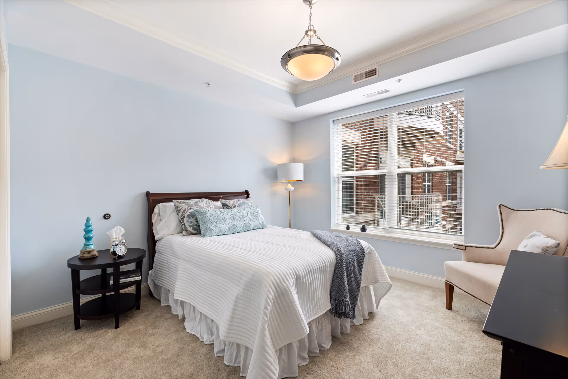 A bright bedroom with light blue walls featuring a neatly made bed with white bedding and decorative pillows. There is a round black bedside table with a blue decorative object, a clock, and a tissue box. A floor lamp stands in the corner next to the bed. A large window with white blinds lets in natural light and shows a view of a brick building outside. A beige armchair with a pillow is positioned near the window, and a black piece of furniture is partially visible on the right side of the image.