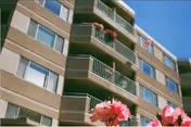 Multi-story beige building with balconies, an American flag, and pink flowers in the foreground against a blue sky.