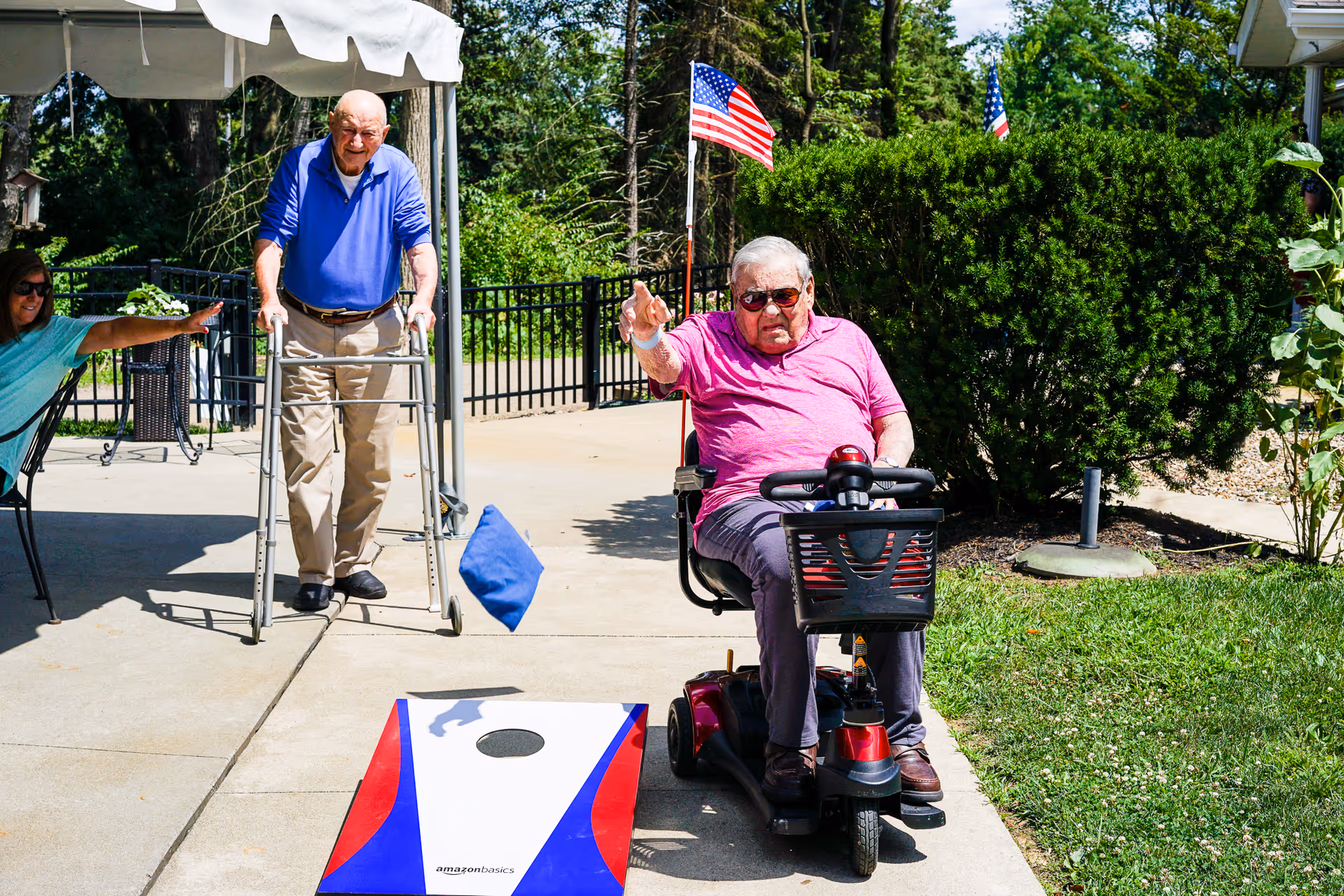 Two elderly men outdoors at a nursing home. One man in a blue shirt uses a walker while playing a bean bag toss game, and another man in a pink shirt and sunglasses sits on a mobility scooter, throwing a bean bag. A woman is seated nearby, watching and cheering. There are American flags and greenery in the background.