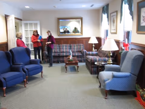 A cozy living room area with several upholstered chairs and sofas arranged around a small wooden coffee table. There are two table lamps on side tables, framed artwork on the walls, and a large mirror reflecting part of the room. Three people are standing and conversing near an open door on the left side of the room.