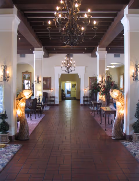 Interior hallway of a senior living facility with a tiled floor, wooden beamed ceiling, and two ornate chandeliers. The hallway is flanked by columns and decorated with elegant furniture, floral arrangements, wall sconces, and two large sculptural lamps shaped like birds.