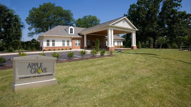 Exterior view of a single-story building with a covered entrance and a sign in the foreground that reads 'Apple Grove Alzheimer's and Dementia Residence'. The building is surrounded by a well-maintained lawn and trees under a clear blue sky.
