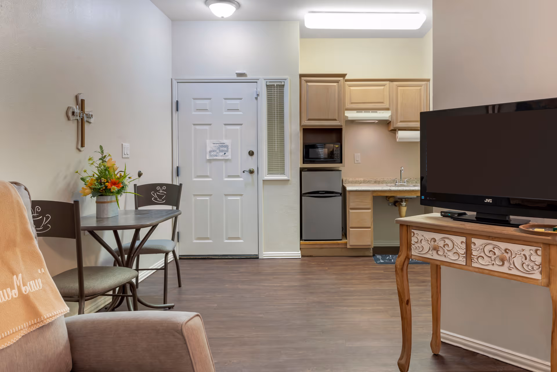 Interior view of a senior living facility room showing a small kitchenette with wooden cabinets, a mini refrigerator, microwave, and sink. To the left is a small round table with two chairs and a flower arrangement. A white door with a small window is in the background. On the right side, there is a wooden table with decorative carvings holding a flat-screen TV. The floor is wooden, and the walls are light-colored.