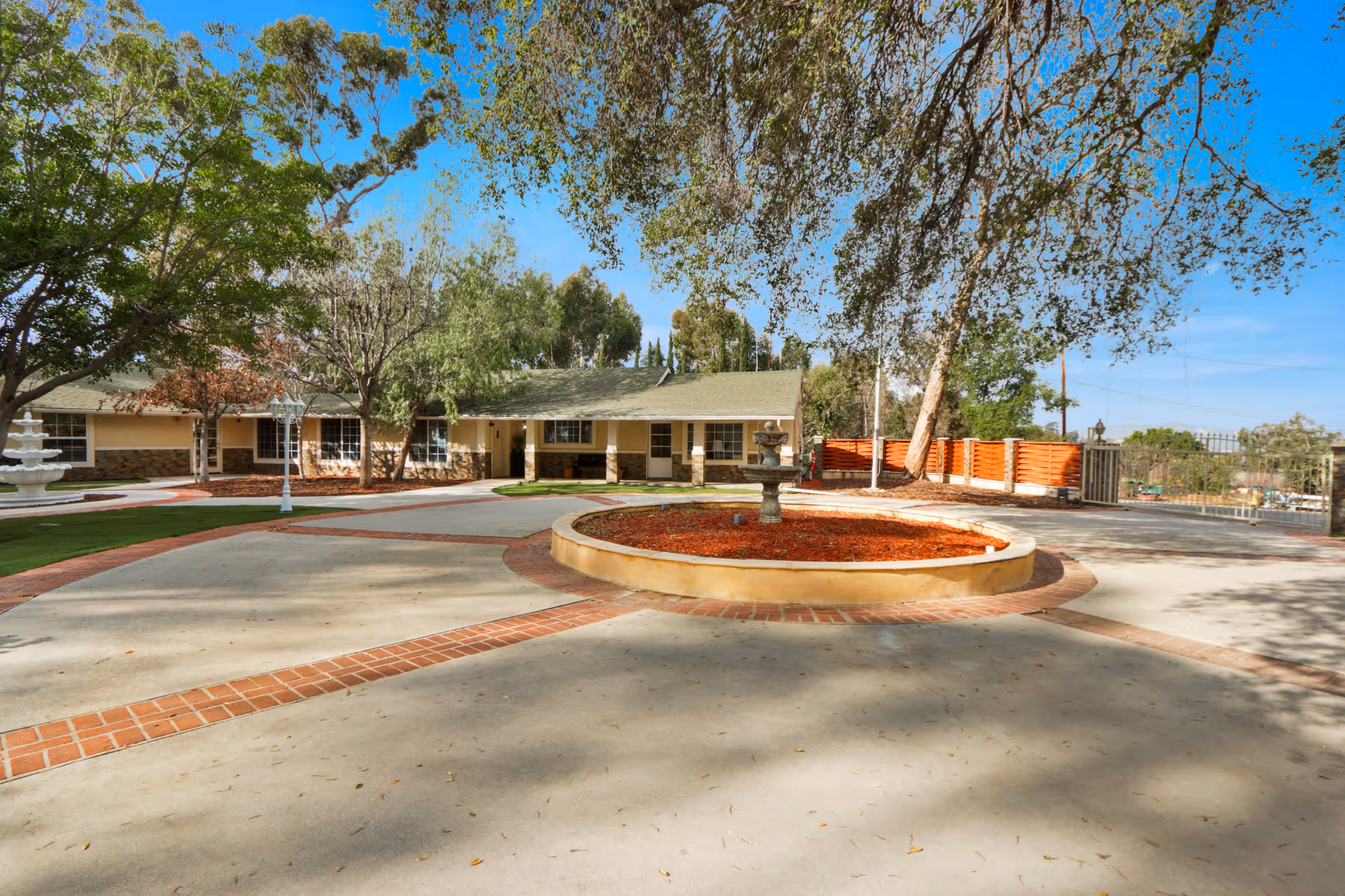 Outdoor view of The Heights Inn showing a circular driveway with a central round planter containing a small fountain. The building is single-story with a green roof and beige walls, surrounded by trees and a clear blue sky.