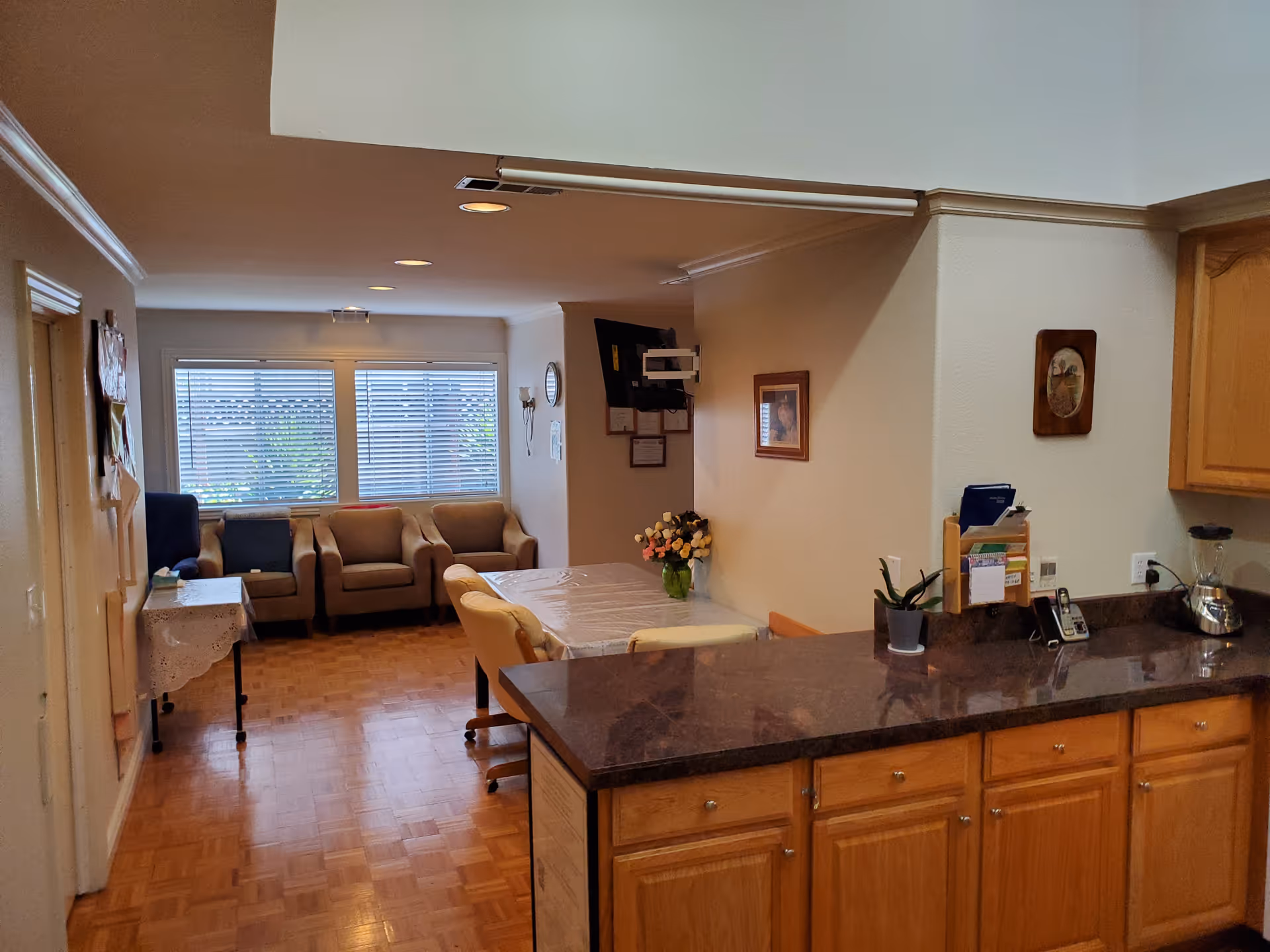 Interior view of a senior living facility common area showing a kitchen counter with wooden cabinets and a dark countertop in the foreground. Behind the counter is a dining table covered with a plastic tablecloth and surrounded by chairs. In the background, there are three armchairs placed near a window with blinds. The walls are decorated with framed pictures and a wall-mounted TV is visible.