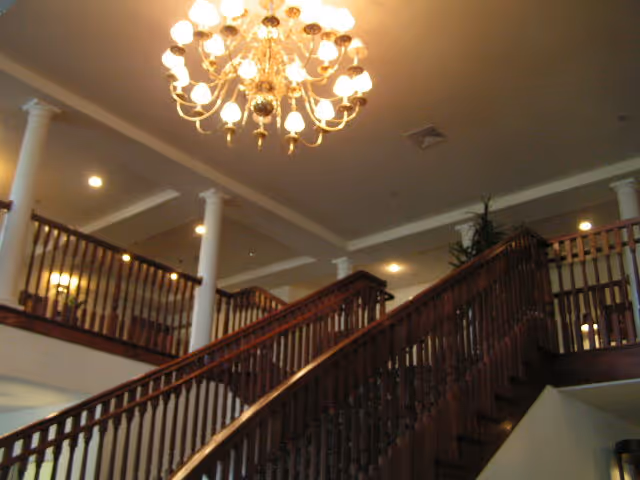 Interior view of a senior living facility showing a grand wooden staircase with ornate railings leading to an upper floor balcony. The ceiling features a large, elegant chandelier with multiple lights, and several white columns support the upper level. The lighting is warm and the space appears spacious and welcoming.